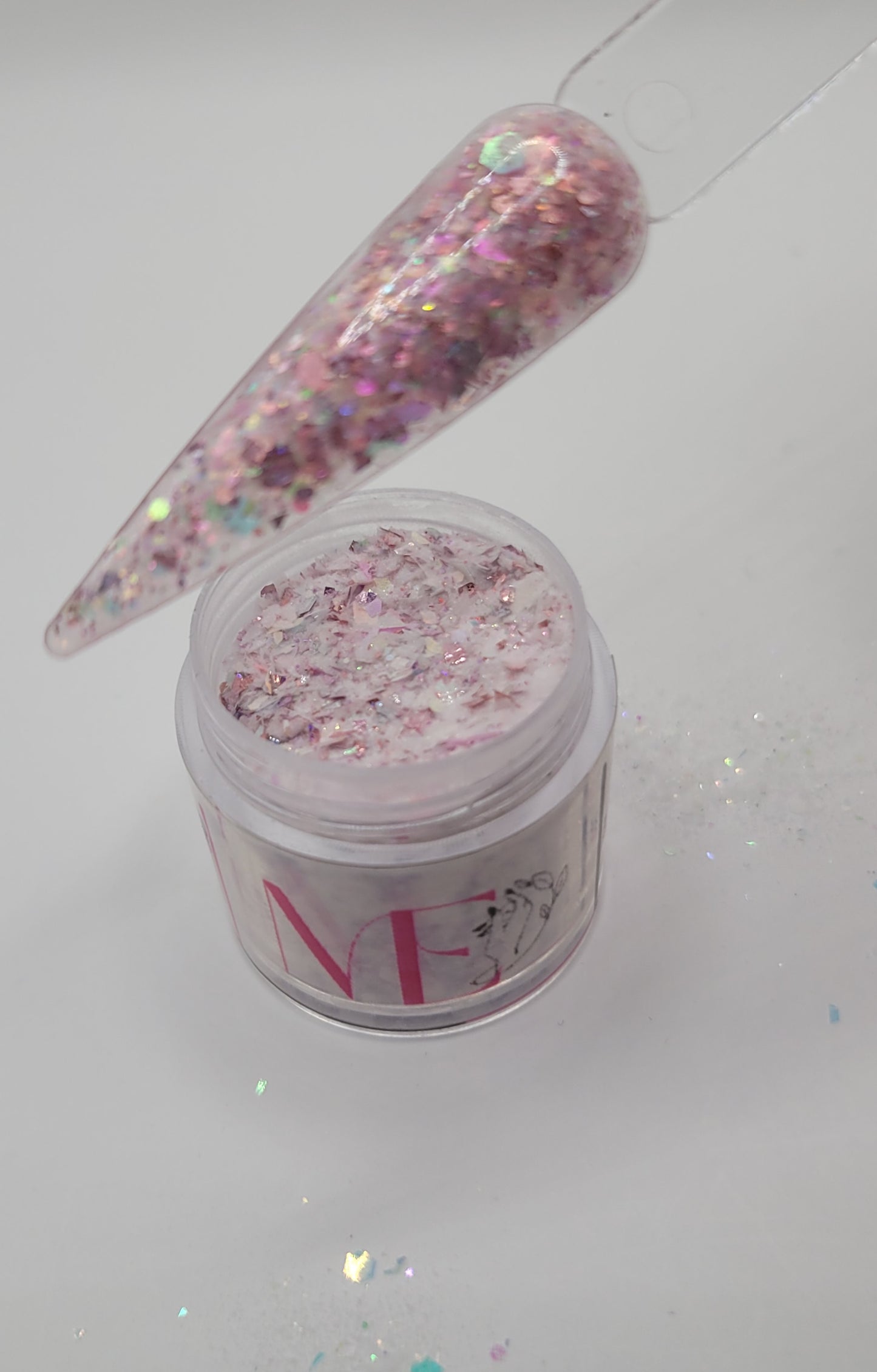 open Jar of pink decorative glitter foil acrylic mix top opened with a swatch stick over top on a white background